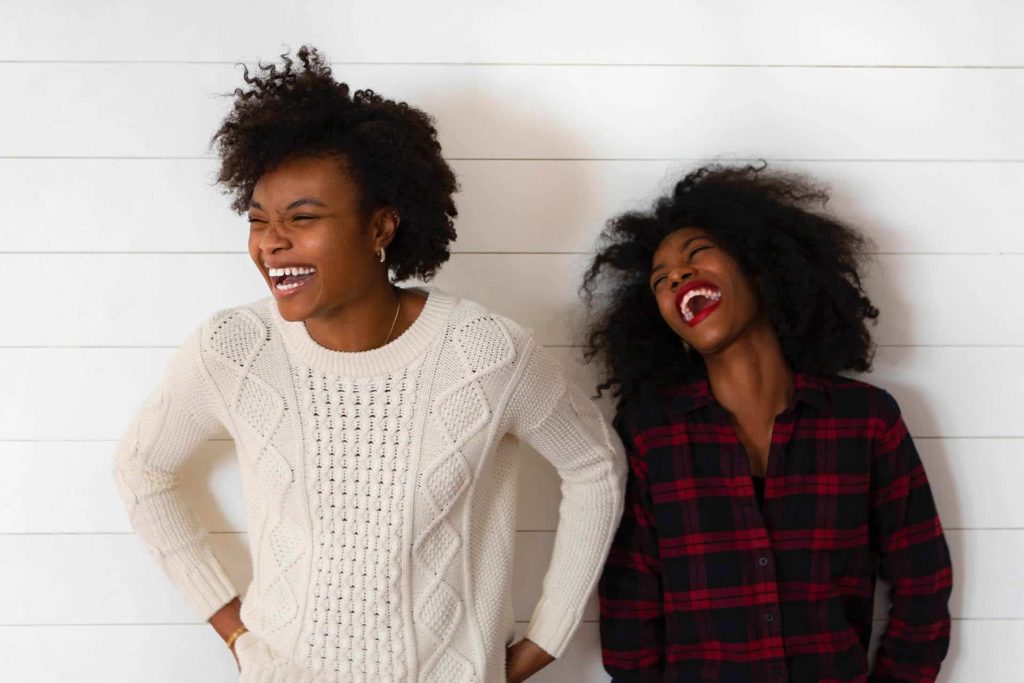 Photo of two black women laughing in front of a white siding background.
