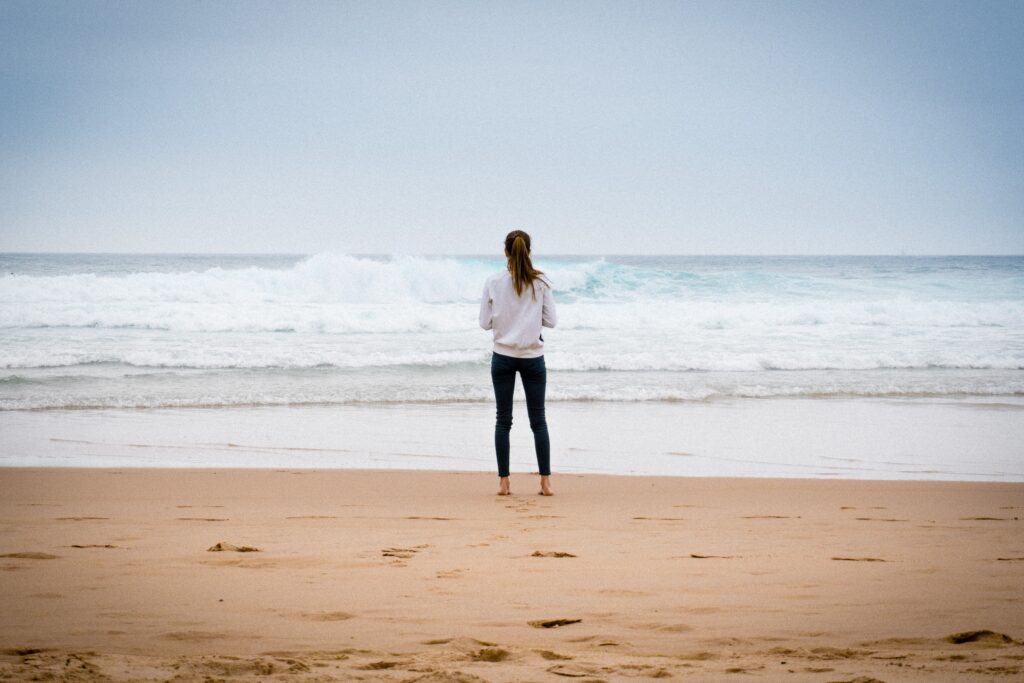 A young woman standing on a beach looking out at the ocean.